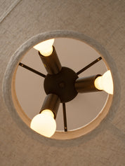 A modern four-light ceiling fixture with a dark central hub and cylindrical arms, each holding a glowing warm white bulb, shown installed on a textured beige ceiling.
