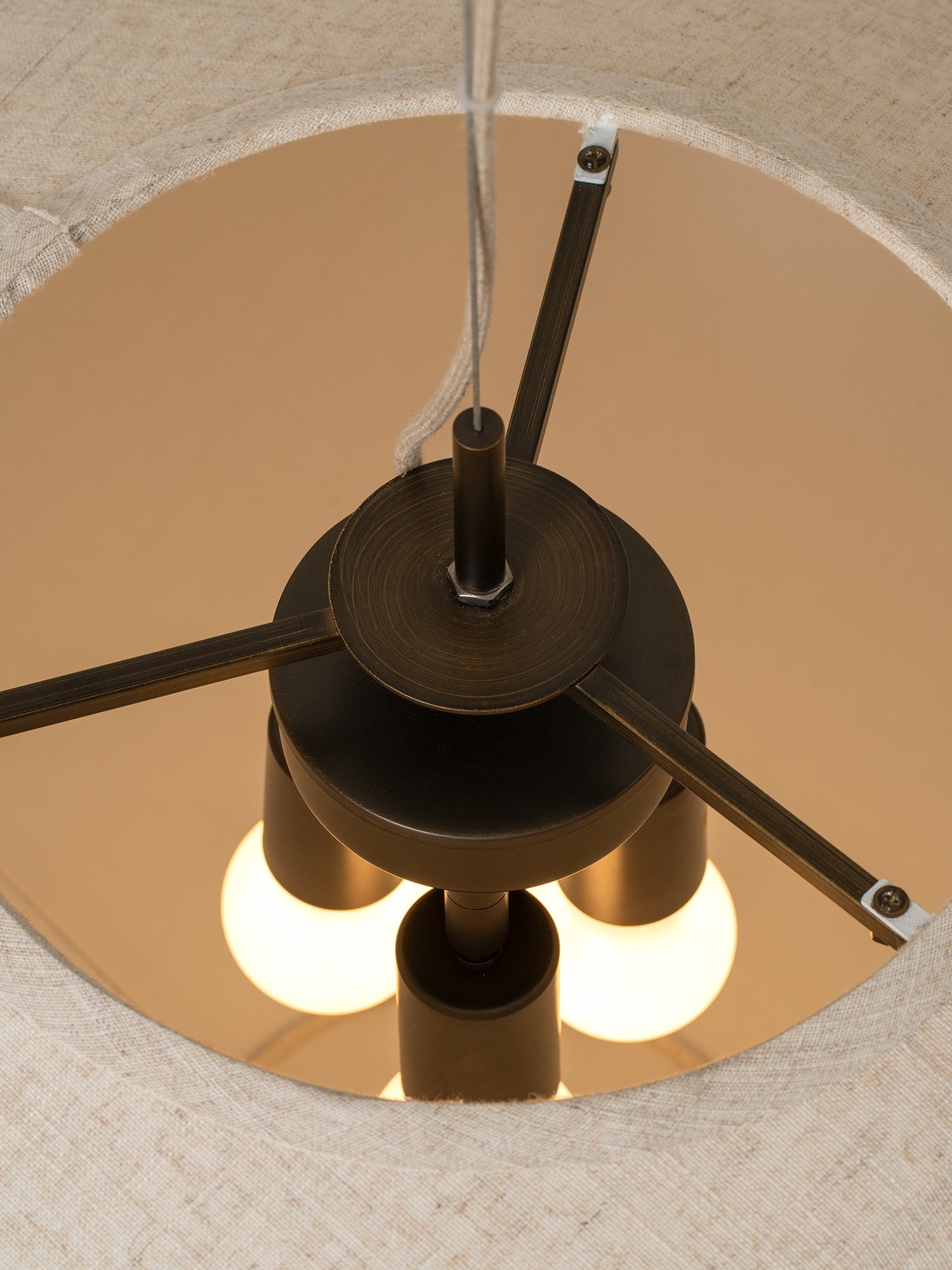 A close-up view from inside a light beige fabric drum shade, showing the top of a three-light ceiling fan with a dark bronze finish and glowing warm white bulbs.