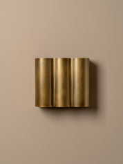 A modern three-cylinder wall sconce in brushed gold metal, mounted on a solid beige wall.