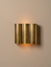 A modern wall-mounted light fixture with three vertical brushed brass cylindrical shades, emitting a warm glow against a solid beige wall.