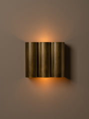 A modern three-cylinder wall sconce in a brushed bronze finish, emitting a warm golden light against a neutral taupe wall.