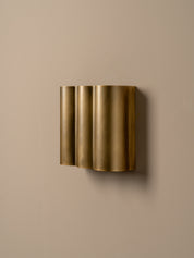 A modern wall-mounted light fixture with three vertically stacked cylindrical shades in a brushed gold finish, mounted on a solid beige wall.
