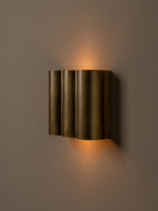 A modern wall-mounted sconce with three vertically fluted cylindrical shades in a brushed brass finish, emitting a warm golden light against a dark taupe wall.