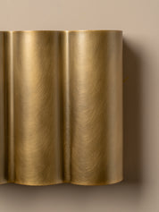 A close-up of a modern wall sconce with a brushed brass or gold metallic finish, featuring a wavy, fluted design mounted on a beige wall.