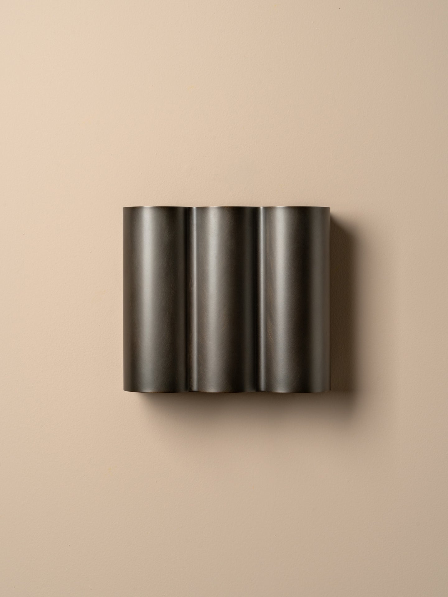 A modern three-cylinder wall sconce in a dark charcoal metallic finish, mounted on a warm beige wall.