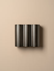 A modern three-cylinder wall sconce in a dark charcoal metallic finish, mounted on a warm beige wall.