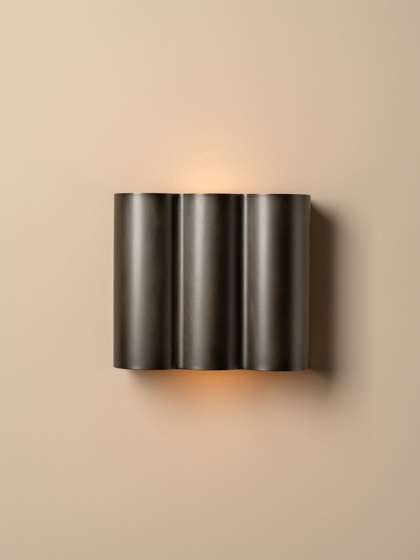 A modern three-cylinder wall sconce in a dark gunmetal finish, mounted on a warm beige wall and casting a soft warm light downward.