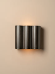 A modern three-cylinder wall sconce in a dark gunmetal finish, mounted on a warm beige wall and casting a soft warm light downward.