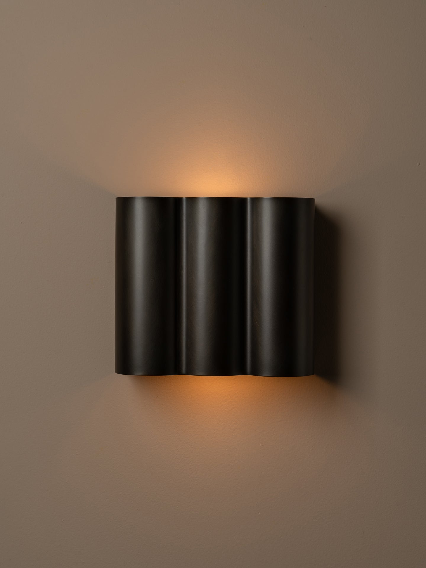 A modern three-cylinder black wall sconce with warm orange light glowing from its top and bottom, mounted on a solid taupe-brown wall.