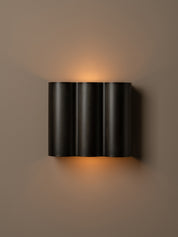 A modern three-cylinder black wall sconce with warm orange light glowing from its top and bottom, mounted on a solid taupe-brown wall.