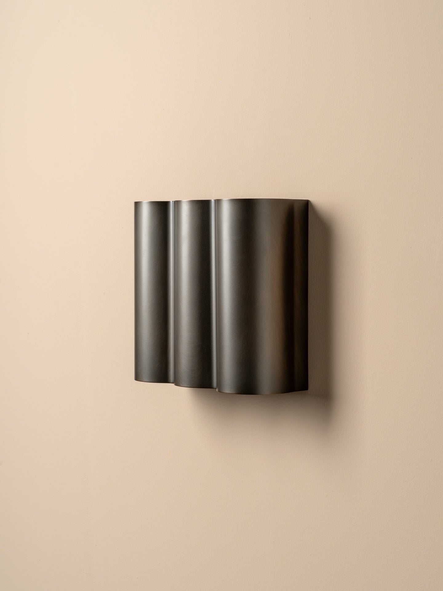 A modern wall-mounted light fixture with three vertically stacked, fluted cylindrical shades in a dark metallic finish, mounted on a plain beige wall.
