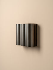 A modern wall-mounted light fixture with three vertically stacked, fluted cylindrical shades in a dark metallic finish, mounted on a plain beige wall.