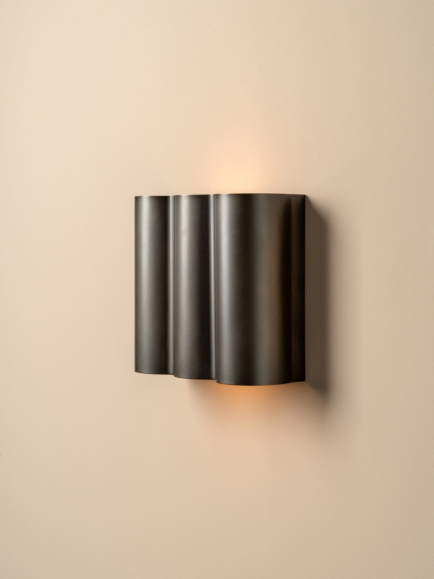 A modern three-cylinder wall sconce in a dark gunmetal finish, mounted on a warm beige wall and casting a soft warm light upwards and downwards.