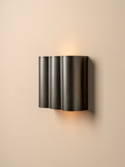 A modern three-cylinder wall sconce in a dark gunmetal finish, mounted on a warm beige wall and casting a soft warm light upwards and downwards.