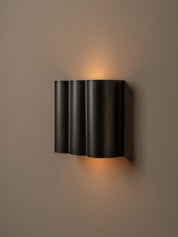 A modern black wall sconce with three vertically fluted cylindrical shades, mounted on a warm taupe wall and casting a soft warm light upward and downward.