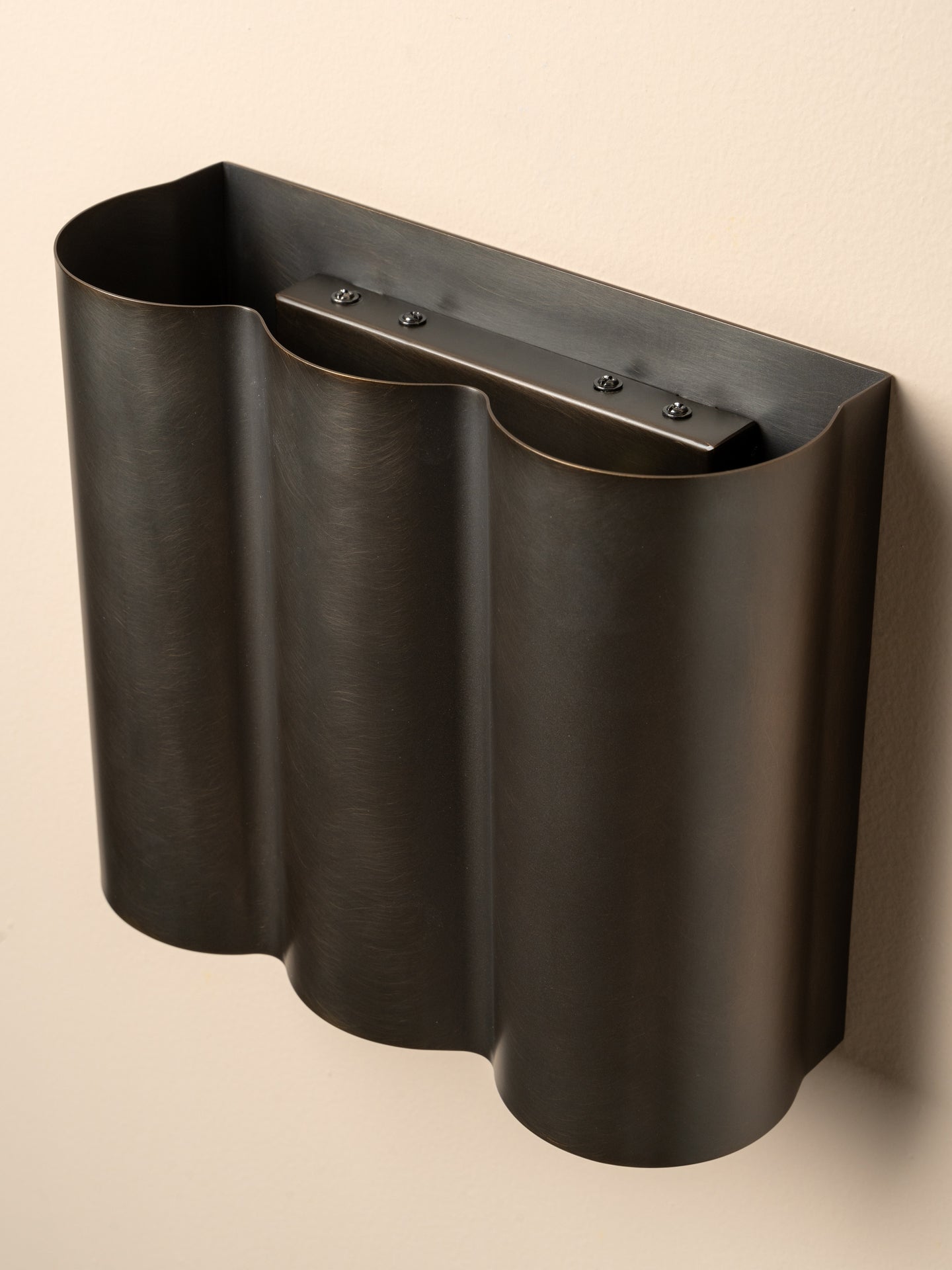 A dark metallic wall-mounted holder with a wavy, fluted silhouette and a horizontal bar with small screws at the top, shown against a plain beige wall.