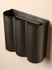 A dark metallic wall-mounted holder with a wavy, fluted silhouette and a horizontal bar with small screws at the top, shown against a plain beige wall.