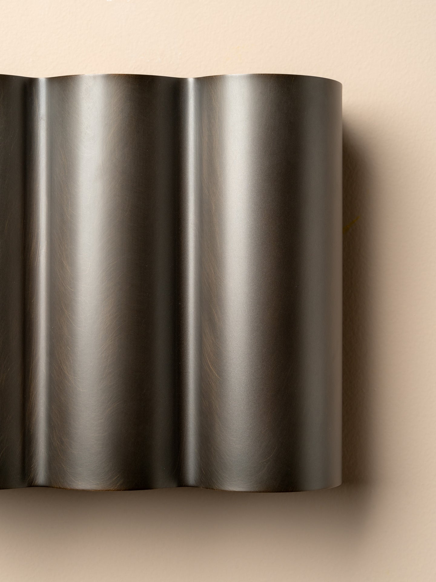 A close-up of a dark metallic, wavy ribbed surface, likely a brushed metal wall sconce or decorative panel, set against a solid beige background.