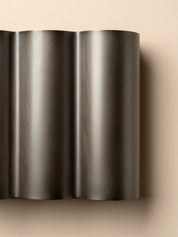 A close-up of a dark metallic, wavy ribbed surface, likely a brushed metal wall sconce or decorative panel, set against a solid beige background.