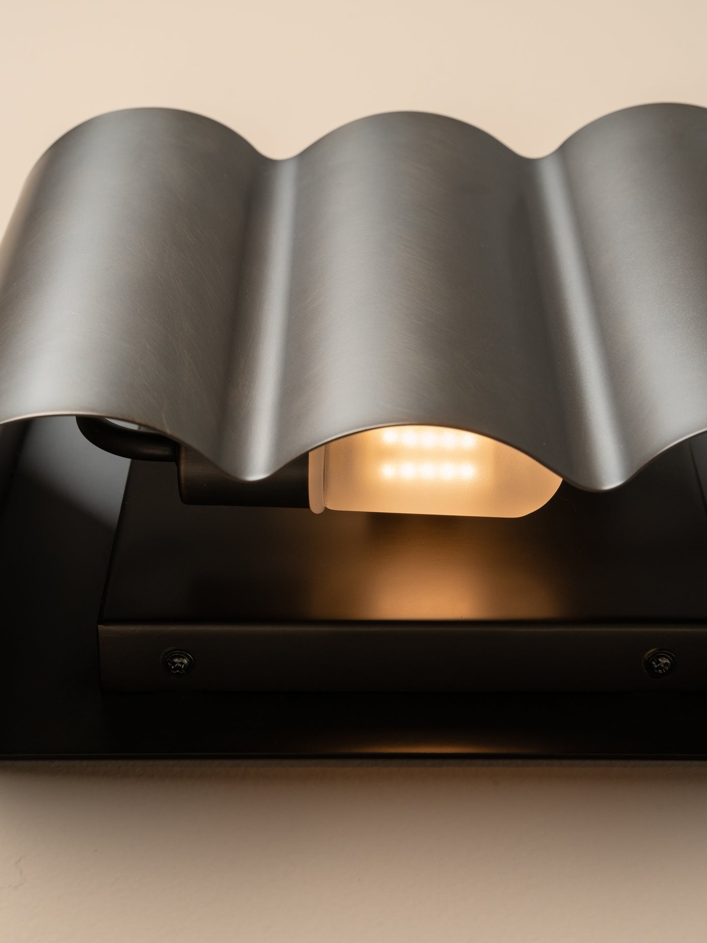 Close-up of a modern wall light fixture with a wavy metallic gray shade and a black base, emitting a warm yellow glow from the illuminated bulb.