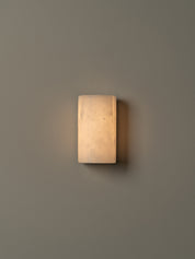 A rectangular wall sconce with a warm, glowing frosted glass shade mounted on a solid gray wall.
