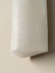 A close-up of a cylindrical wall sconce with a frosted white glass shade mounted on a beige textured wall.
