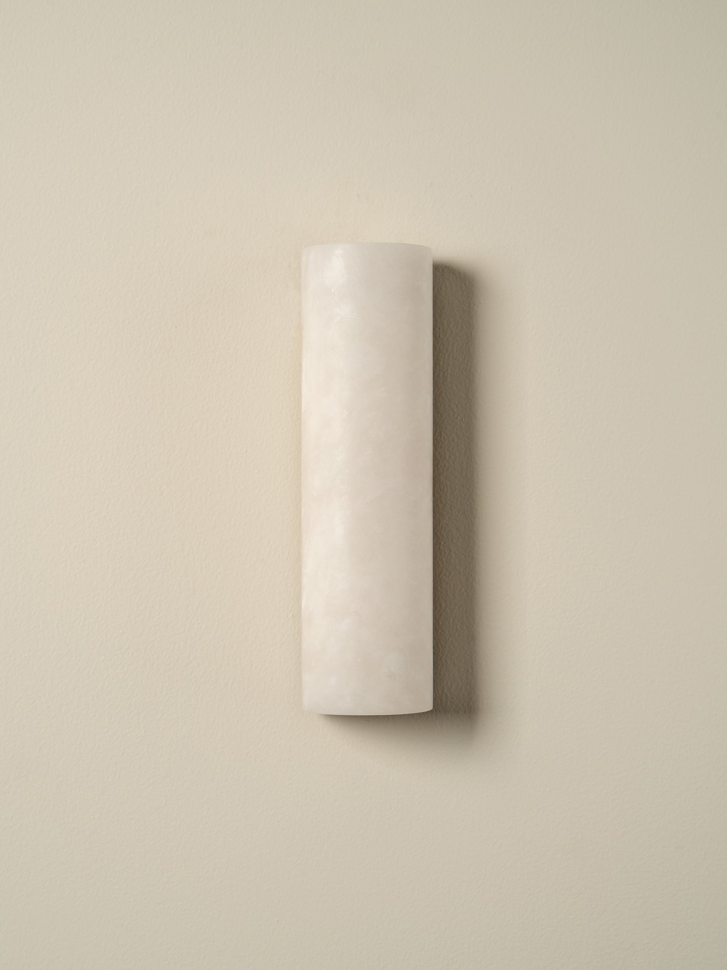 A minimalist cylindrical wall sconce in a textured off-white finish, mounted on a solid beige wall with a soft shadow.