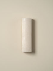 A minimalist cylindrical wall sconce in a textured off-white finish, mounted on a solid beige wall with a soft shadow.
