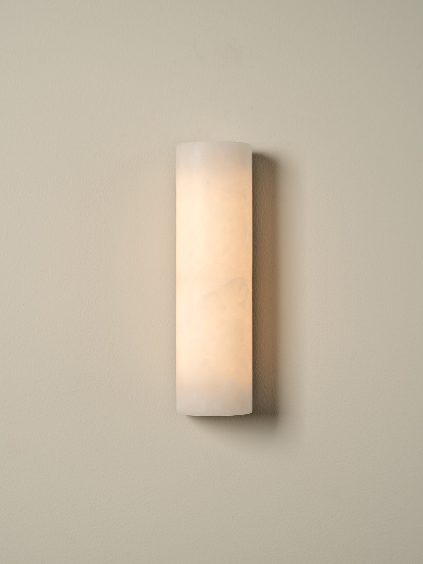 A modern cylindrical wall sconce with a frosted white glass shade, emitting a warm light against a neutral beige wall.