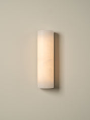 A modern cylindrical wall sconce with a frosted white glass shade, emitting a warm light against a neutral beige wall.