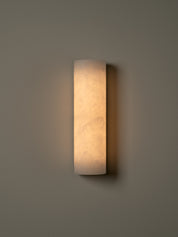 A modern cylindrical wall sconce with a warm glowing frosted glass shade, mounted on a neutral gray wall.