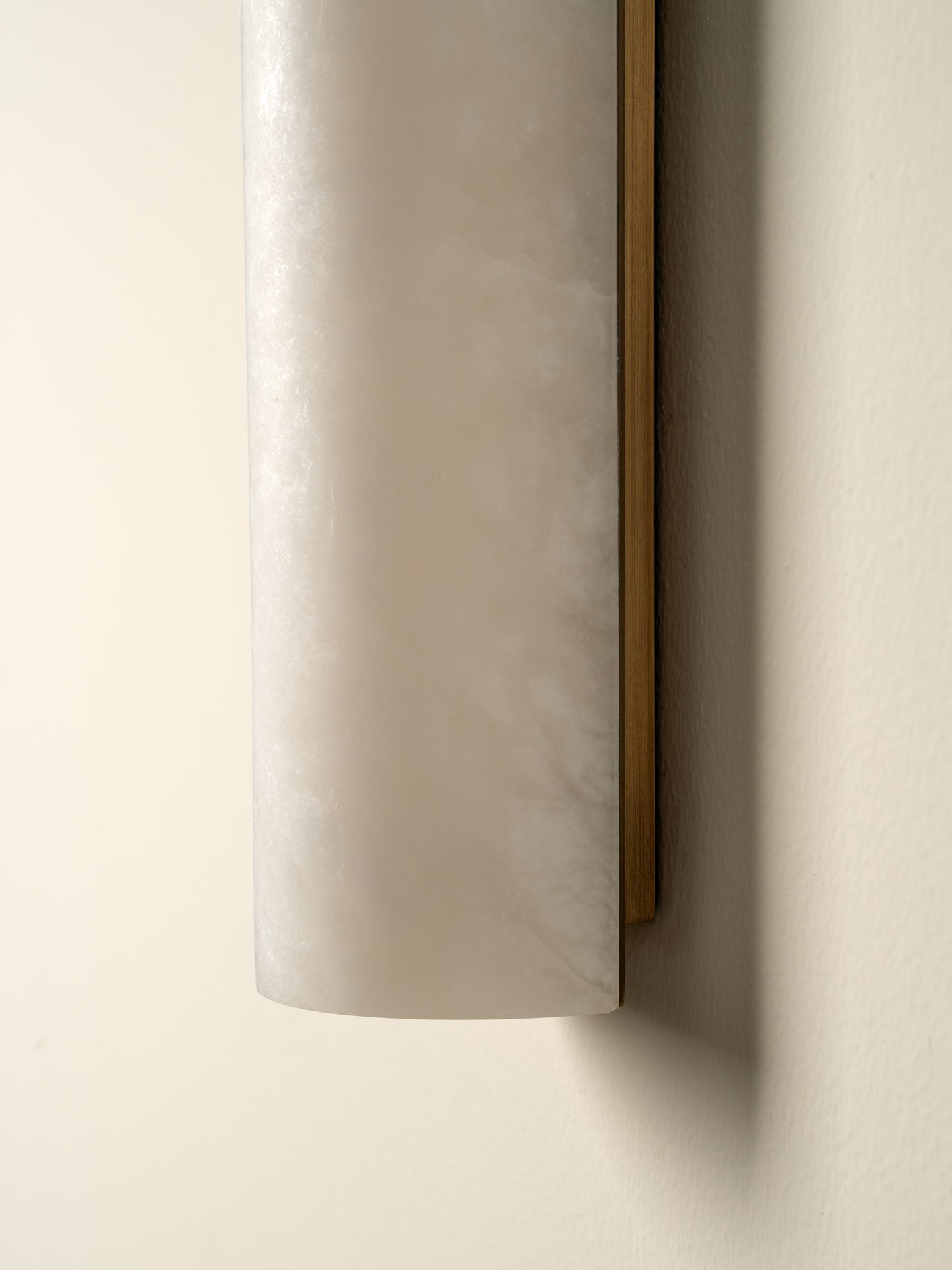 A close-up of a cylindrical wall sconce with a frosted white marble or stone shade and a slim gold-toned side frame, mounted on a warm beige wall.