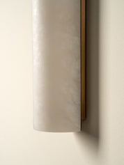 A close-up of a cylindrical wall sconce with a frosted white marble or stone shade and a slim gold-toned side frame, mounted on a warm beige wall.