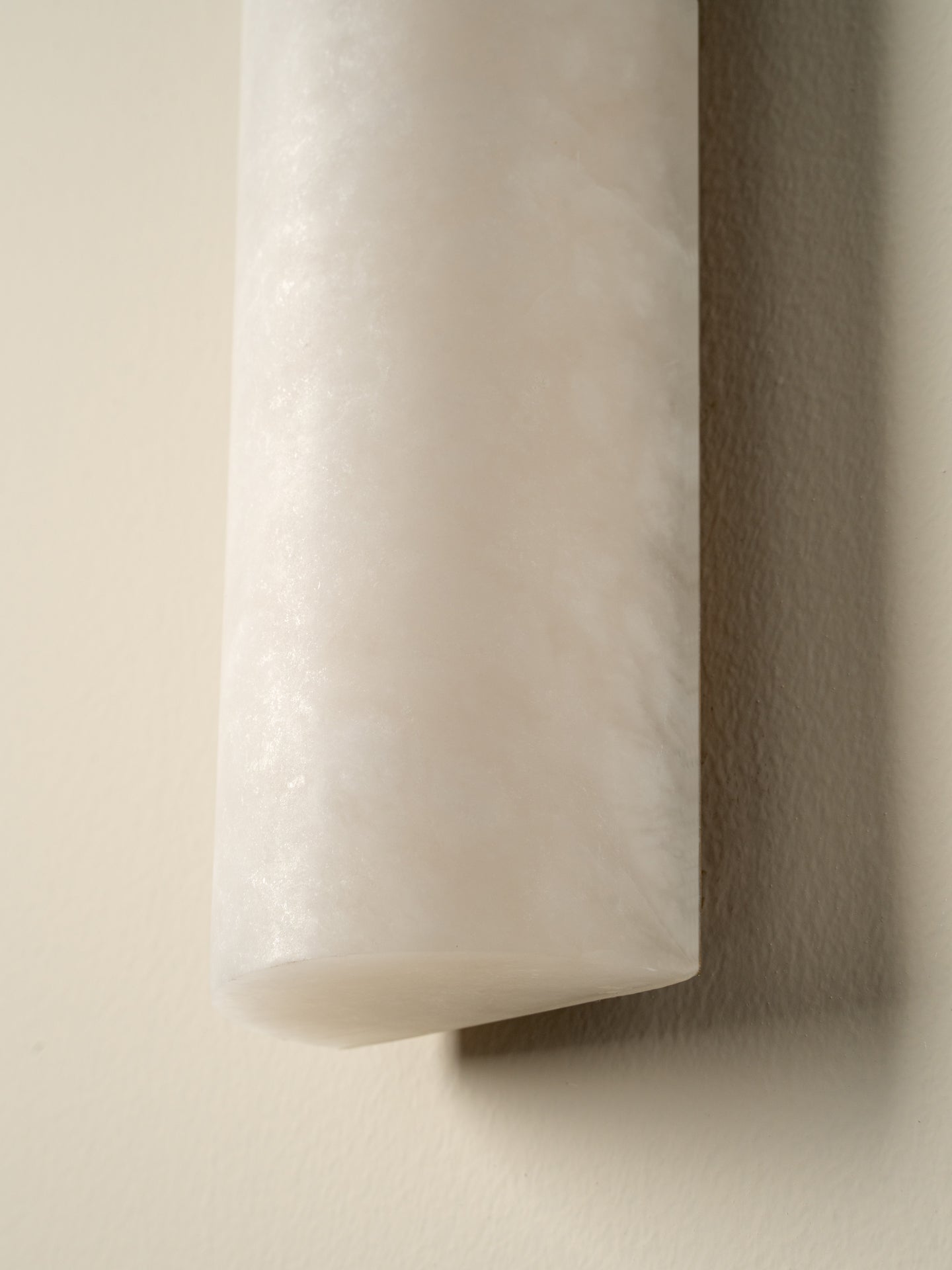 A close-up of a cylindrical wall sconce with a frosted, textured white material, mounted on a warm beige wall casting a soft shadow.