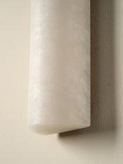 A close-up of a cylindrical wall sconce with a frosted, textured white material, mounted on a warm beige wall casting a soft shadow.