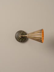 A modern wall-mounted lamp with a fluted brass shade, flexible curved neck, and a round dark green and brown stone backplate, shown against a plain beige wall.