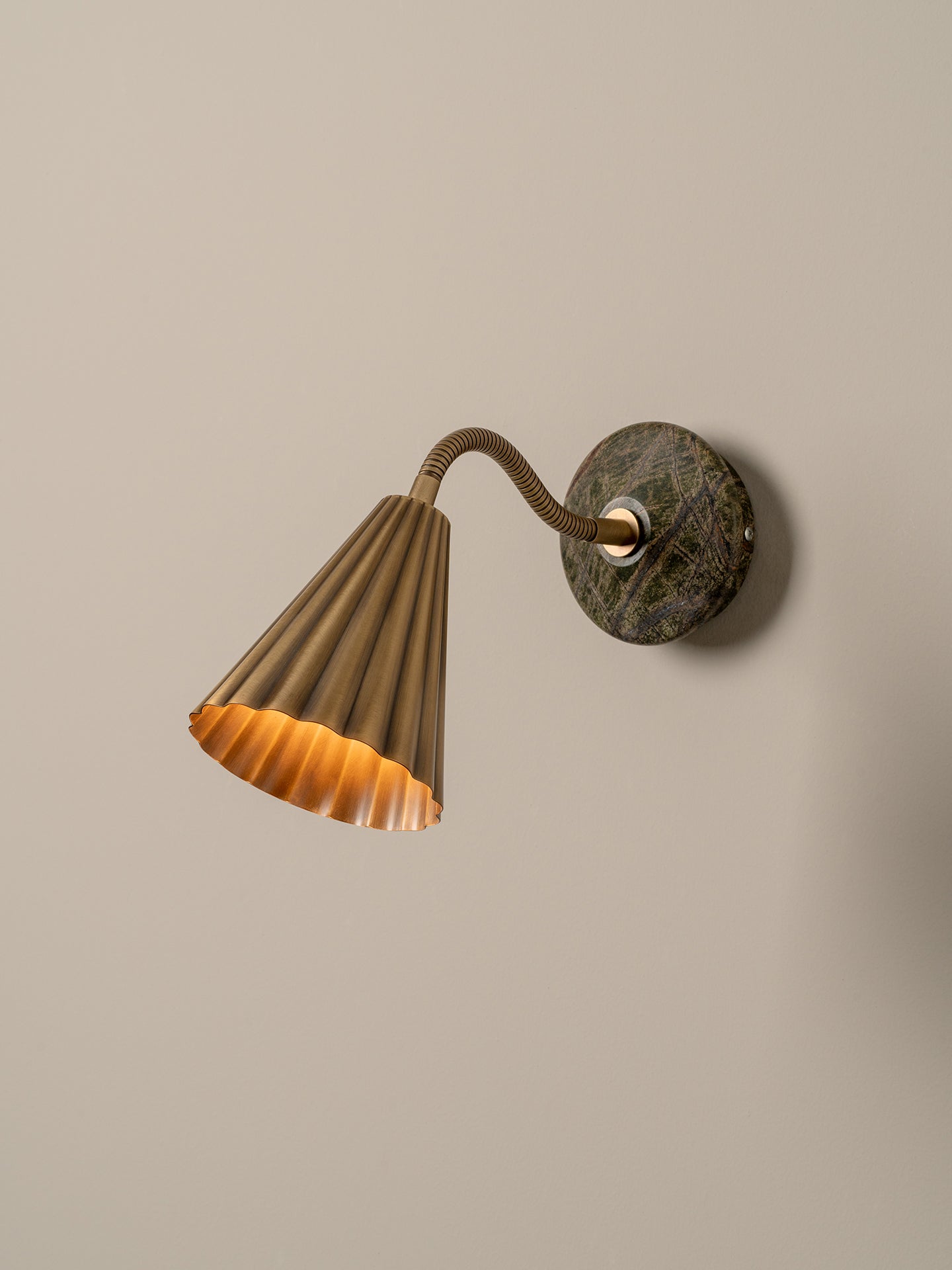 A modern wall-mounted lamp with a fluted brass shade, flexible curved neck, and a round dark green and brown stone backplate, shown against a plain beige wall.