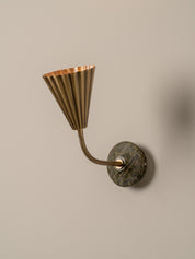 A modern wall-mounted lamp with a fluted brass shade, flexible curved neck, and a round dark green and brown stone backplate, shown against a plain beige wall.