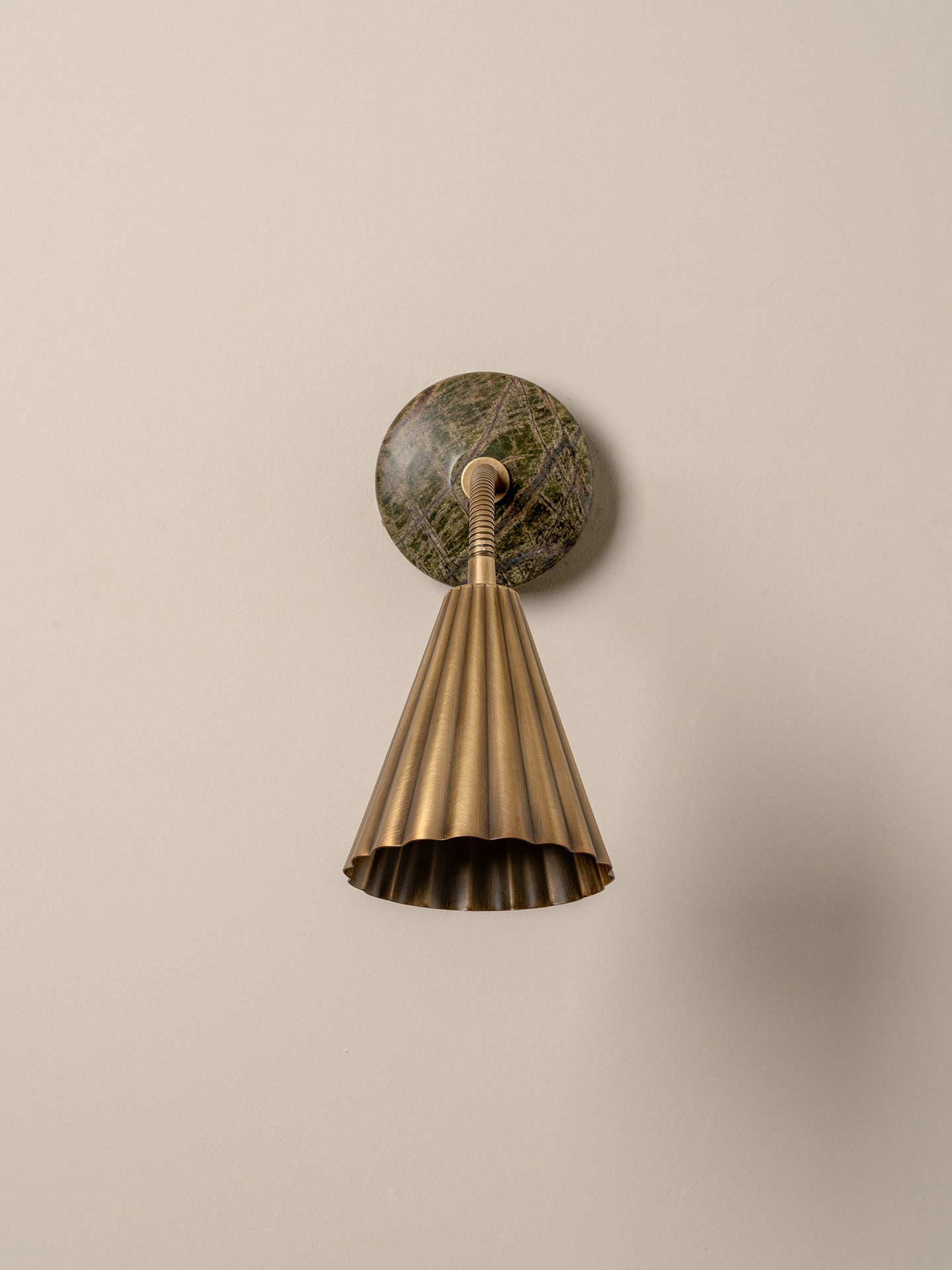 A modern wall-mounted lamp with a fluted brass shade, flexible curved neck, and a round dark green and brown stone backplate, shown against a plain beige wall.