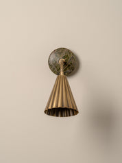 A modern wall-mounted lamp with a fluted brass shade, flexible curved neck, and a round dark green and brown stone backplate, shown against a plain beige wall.