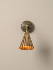 A modern wall sconce light fixture featuring a fluted brass cone shade and a round green marble wall mount, shown against a plain beige wall.