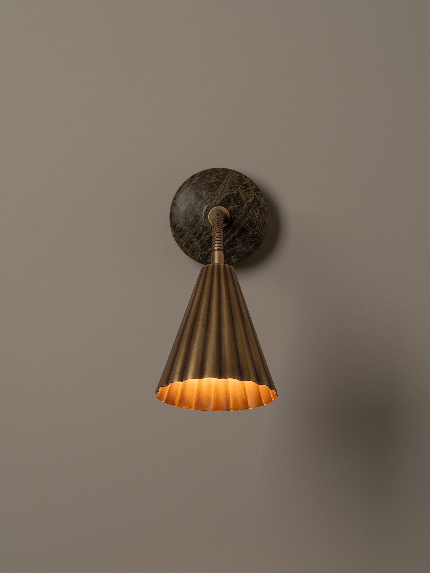 A modern wall-mounted lamp with a fluted brass shade, flexible curved neck, and a round dark green and brown stone backplate, shown against a plain beige wall.