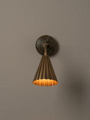 A modern wall-mounted lamp with a fluted brass shade, flexible curved neck, and a round dark green and brown stone backplate, shown against a plain beige wall.
