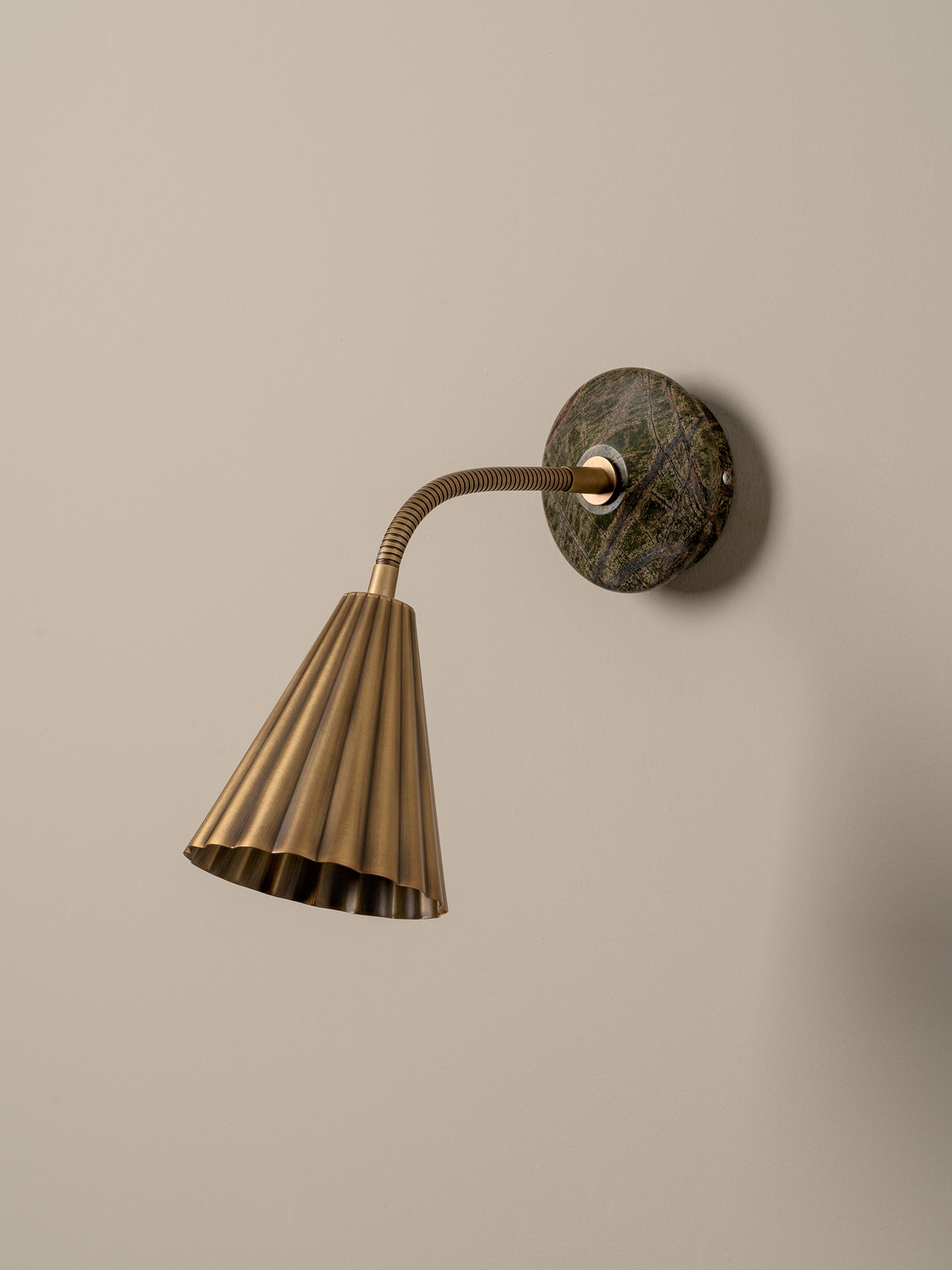 A modern wall-mounted lamp with a fluted brass shade, flexible curved neck, and a round dark green and brown stone backplate, shown against a plain beige wall.