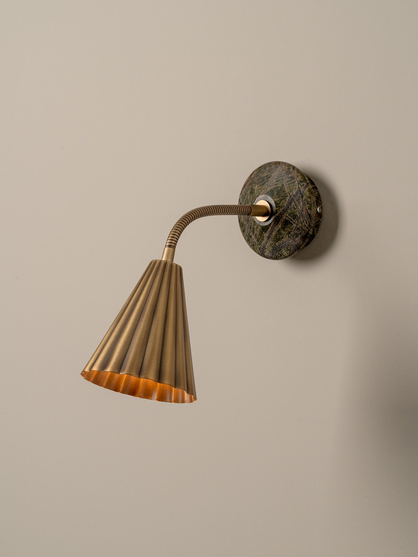 A modern wall-mounted lamp with a fluted brass shade, flexible curved neck, and a round dark green and brown stone backplate, shown against a plain beige wall.