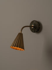 A modern wall-mounted lamp with a fluted brass shade, flexible curved neck, and a round dark green and brown stone backplate, shown against a plain beige wall.