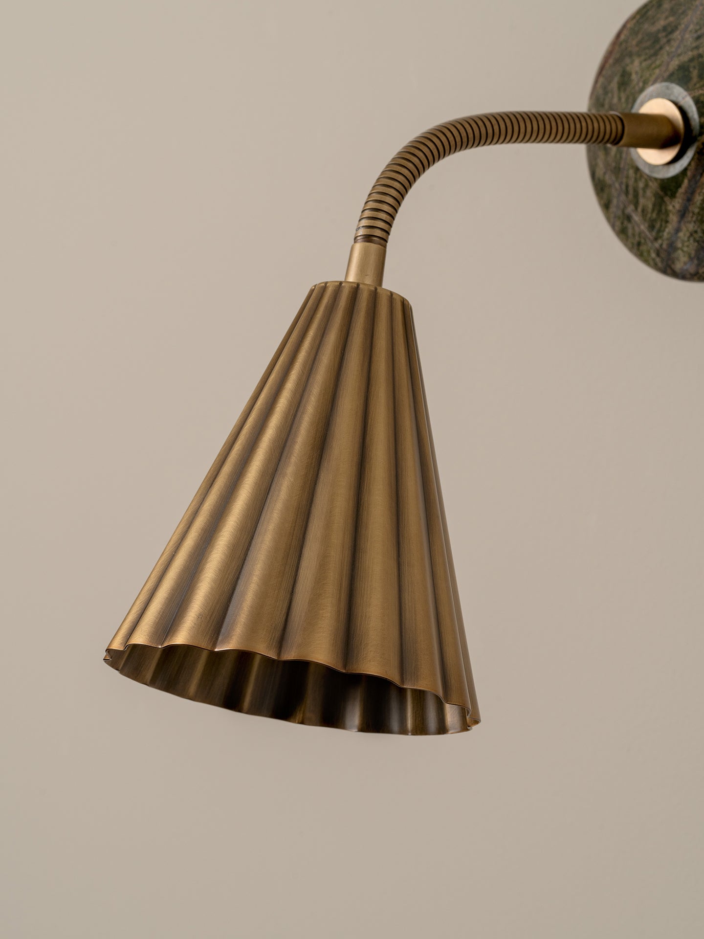 A close-up of a modern wall sconce with a fluted bronze metal shade and a flexible curved arm, mounted against a plain beige wall.