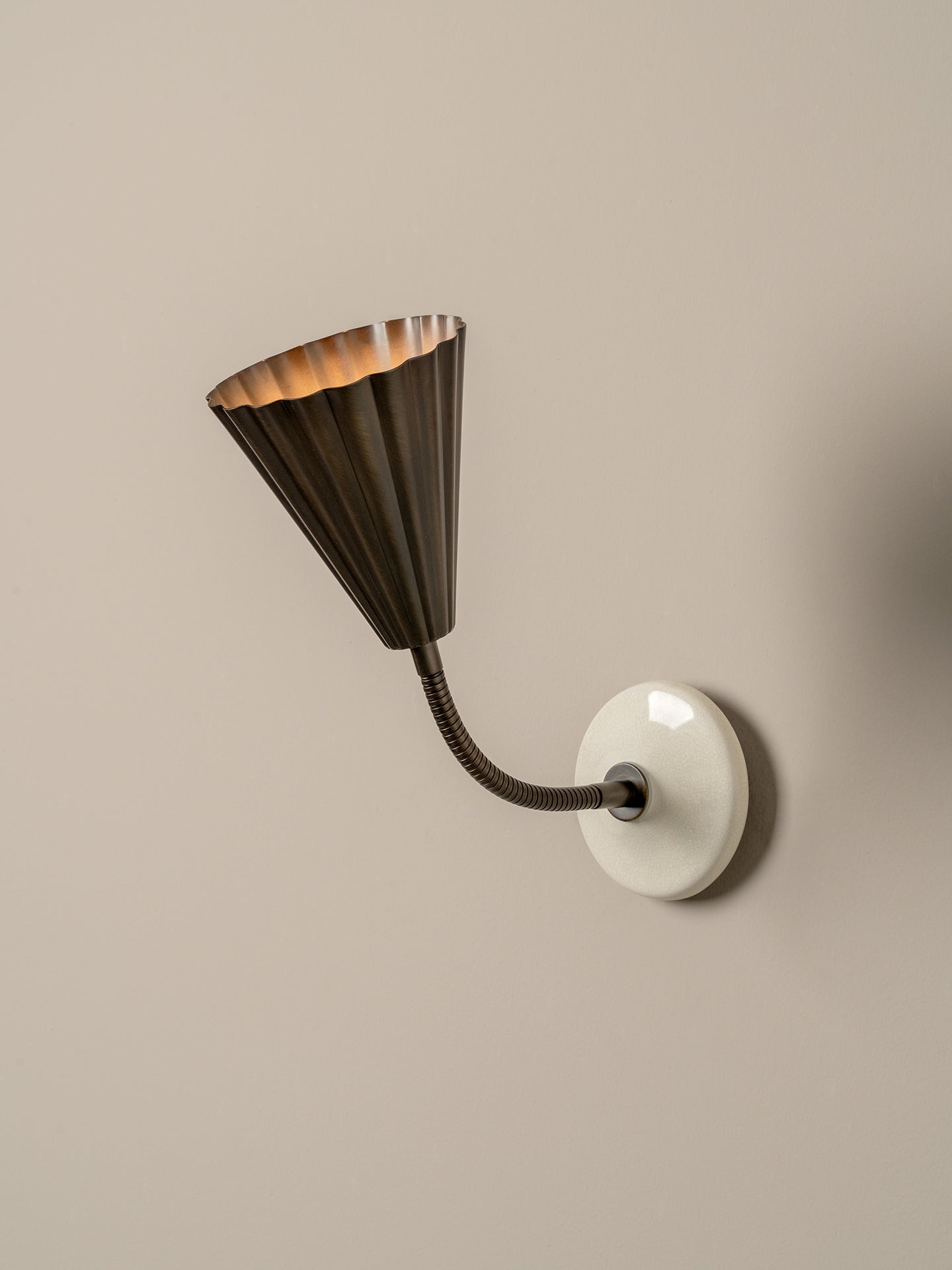 A modern wall sconce with a dark bronze fluted conical shade and a flexible curved arm, mounted on a round white wall plate against a warm beige wall.