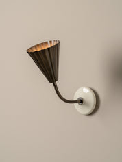 A modern wall sconce with a dark bronze fluted conical shade and a flexible curved arm, mounted on a round white wall plate against a warm beige wall.