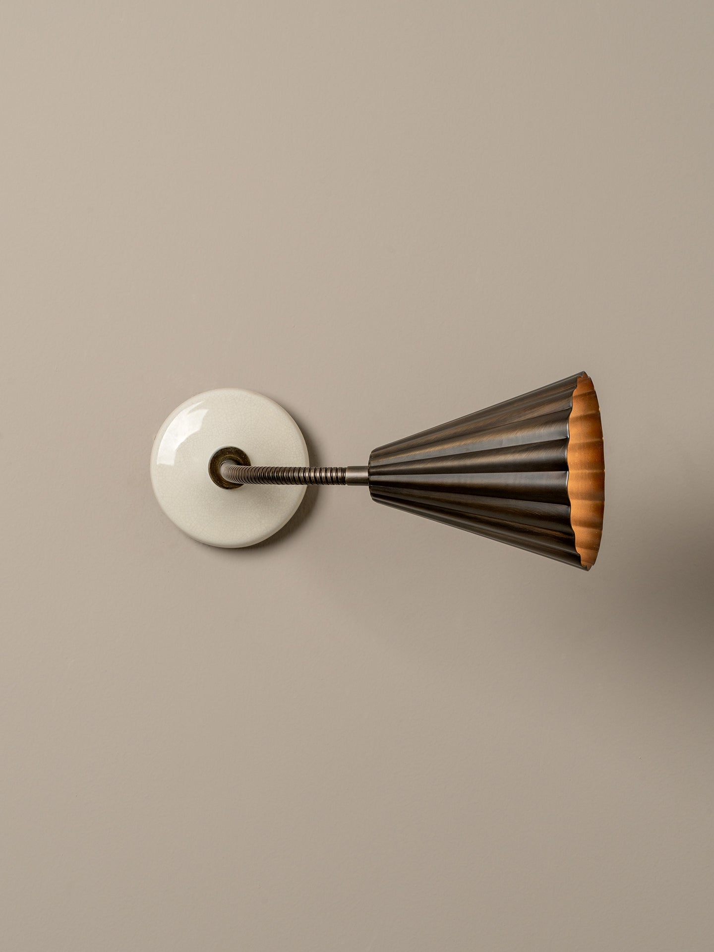 A modern wall sconce with a circular white backplate, a dark metal adjustable arm, and a flared shade featuring a dark bronze exterior and a warm orange-gold interior, mounted on a solid beige wall.